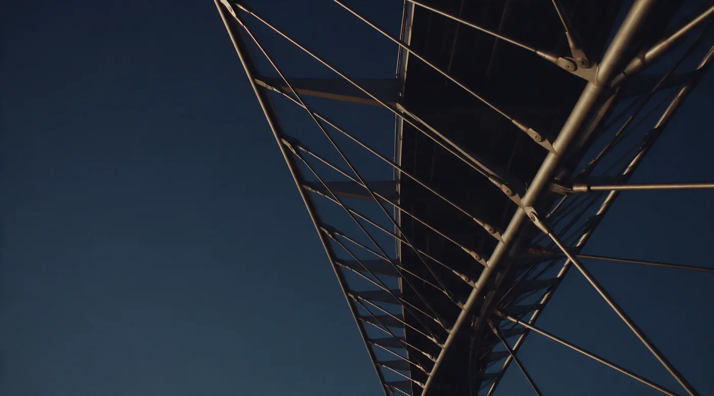 Structure d’une passerelle métallique vue d’en dessous au crépuscule, câbles éclairés d’une lumière dorée.